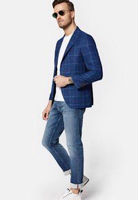 Niebieska marynarka w kratę z kontrastującymi czerwonymi liniami, wykonana z teksturowanej tkaniny. Biały t-shirt i wyblakłe dżinsy, dopełnione białymi trampkami.