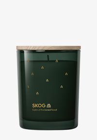 Mörkgrön glasljus med trälock, små gyllene trädkonstikoner och texten "SKOG Lugnet i den boreala skogen" på framsidan.