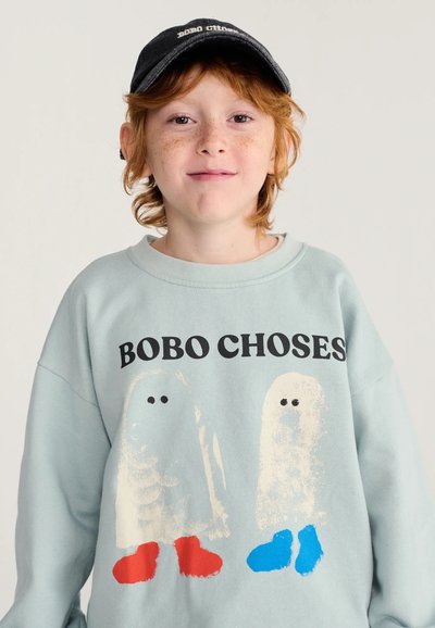 Bambino dai capelli rossi che indossa un berretto nero e una felpa azzurro chiaro con due figure di fantasmi e il testo "BOBO CHOSES" su uno sfondo neutro.
