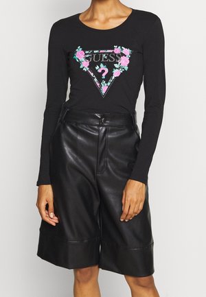 Persoon draagt een zwarte top met lange mouwen met een bloemrijk Guess-logo en zwarte knie-lange shorts van hoogwaardig leer.