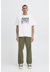 Hvid bomuld t-shirt med et grafisk tryk, kombineret med olivengrønne bukser i løs pasform. Fodtøjet har et sort og hvidt design med gule accenter.