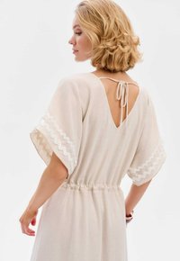 Beige jurk met korte, zwierige mouwen en een verlaagde taille, met een diepe V-rug en witte geschulpte afwerking langs de mouwen.