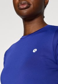 Kék póló sima textúrával és rövid ujjakkal. Bal oldalon egy kis fehér logó található.