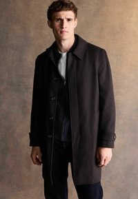 Czarny wełniany płaszcz z kołnierzem, jednorzędowe zapięcie na guziki i ukośne boczne kieszenie. Nosi się go na ciemnym swetrze i dżinsach.