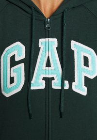 Felpa verde con zip, caratterizzata da una grande scritta "G.A.P." in bianco e turchese, coulisse con punte metalliche e una morbida texture del tessuto.