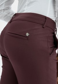 Bordeauxkleurige broek met een gladde textuur, voorzien van een vlakke zak en een zilveren metalen accent. Close-up van de achterkant.