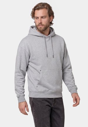 Mand med skæg iført en lysegrå hættetrøje og mørke bukser, kigger ned og til siden mod en ensfarvet baggrund.