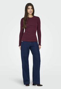 Maglietta lunga color mogano con scollatura ondulata, abbinata a jeans blu scuro a vita alta e gamba larga. Il modello è in piedi con le mani sui fianchi.