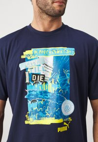 Tumši zils kokvilnas T-krekls ar grafisku izdruku zilās un dzeltenās krāsās, tekstu "MANCHESTER CITY" un apakšā apaļo logotipu.