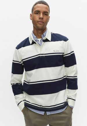 Marinblå och vit randig långärmad pikétröja med knappkrage. Har en logotyp på bröstet och är lager-på-lager med en blå- och vitrandig skjorta under.
