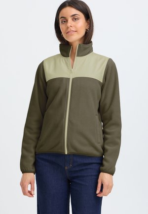 Femme portant une veste polaire vert olive avec des panneaux beiges sur la poitrine et un jean bleu, debout devant un fond clair uni.