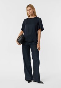 Granatowa bluzka z krótkim rękawem, w połączeniu z ciemnymi dżinsami z rozszerzanymi nogawkami. Modelka trzyma małą czarną torebkę. Prosty, nowoczesny design z gładkimi fakturami.