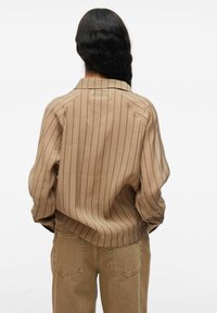 Persona con lunghi capelli ondulati scuri, che indossa una camicia a righe beige larga e pantaloni marroni chiari, vista da dietro su uno sfondo bianco.