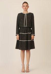 Vestido de tweed negro con una chaqueta corta, adornada con acentos plateados y líneas texturizadas, con mangas largas y una falda acampanada.