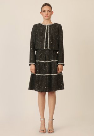 Schwarzes Tweedkleid mit einer kurzen Jacke, verziert mit silbernen Akzenten und strukturierten Linien, mit langen Ärmeln und einem ausgestellten Rock.