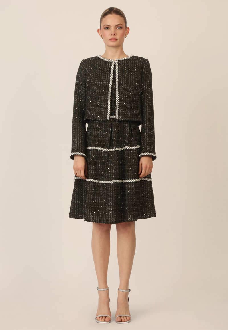 Vestido de tweed negro con una chaqueta corta, adornada con acentos plateados y líneas texturizadas, con mangas largas y una falda acampanada.