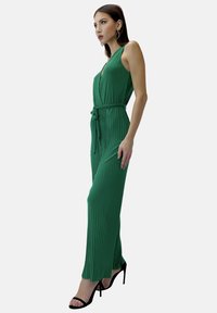 Mono plisado verde con escote en V, con cintura atada, presenta una silueta recta y aberturas laterales, combinado con sandalias de tacón negro.