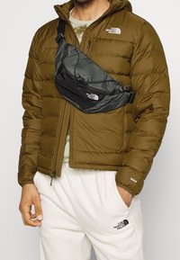 Black nylon crossbody bag worn over an olive green puffer jacket. Distinctive logos visible on both bag and jacket. Soft, quilted texture.