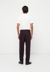 Man standing facing a white wall, wearing a plain white T-shirt, dark purple pants, and black shoes.