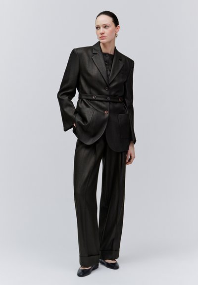 Frau mit dunklen Haaren in einem eleganten schwarzen Blazer mit Gürtel und passenden weiten Hosen, stehend vor einem neutralen grauen Hintergrund.