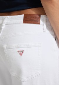 Baltos džinsai su odiniu Guess etiketė ant juosmens ir raudonu bei baltu trikampiu logotipo pėdos užpakalinėje kišenėje.