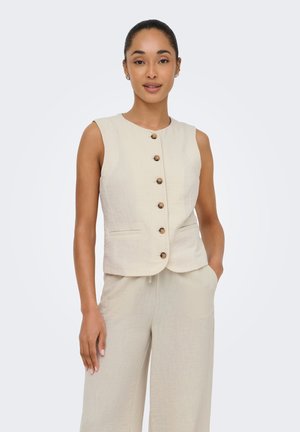 Kvinde iført beige ærmeløs knappet vest og matchende bukser, stående med den ene hånd i lommen mod en ensfarvet baggrund.