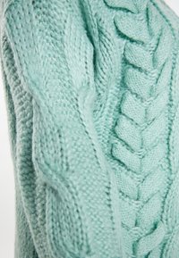Ljus mintgrön handstickad tröja med en texturerad kabeldesign. Har tjock garn och ett mysigt utseende, som visar detaljerade stickmönster.
