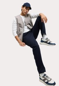 Model trägt eine hellgraue gestreifte Weste über einem weißen Hemd, kombiniert mit marineblauen Hosen und blaugrauen Sneakers mit Akzenten und weißen Sohlen.
