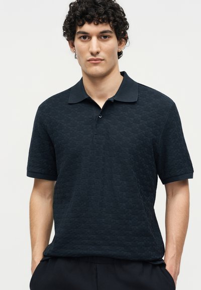 Νέος άνδρας με σγουρά σκούρα μαλλιά, φοράει μαύρο πόλο με υφή και κοντά μανίκια, στέκεται με τα χέρια στις τσέπες, ουδέτερο φόντο.