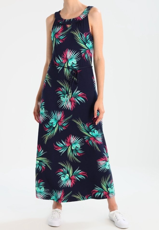 Femme portant une robe maxi sans manches bleu marine avec un imprimé de feuilles tropicales colorées et des baskets blanches.