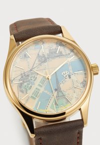 Montre dorée avec un cadran au thème de la carte, présentant des couleurs bleu et beige. Bracelet en cuir brun avec une surface texturée et des détails de couture subtils.