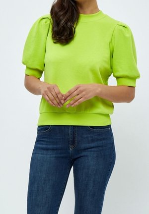 Vrouw draagt een felgroene, korte puffmouwen top en blauwe spijkerbroek, staand met handen zachtjes gevouwen voor zich.