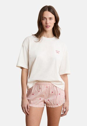 Frau trägt ein weißes übergroßes T-Shirt mit rotem Text und gestreifte Shorts mit kleinen Herzmustern, steht vor einem einfarbigen Hintergrund.