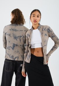 Ribbet zip-up jakke i beige med blå tie-dye mønster. Har høy krage og lange ermer. Båret over en hvit crop top og svart skjørt.