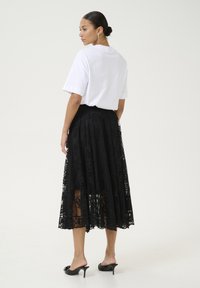 Gonna maxi in pizzo nero con sovrapposizione trasparente, abbinata a una maglietta bianca a maniche corte. Presenta una silhouette dritta e décolleté neri con cui indossarla.