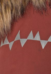 Rød stof med et sølvfarvet zigzag-mønster langs bunden. Blød faux fur-kant øverst tilføjer struktur og visuel kontrast.