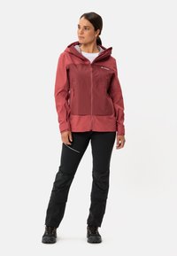 Giacca softshell rossa con cappuccio, frontale con zip e tasche laterali; abbinata a pantaloni neri con dettagli testurizzati e zip alle caviglie.