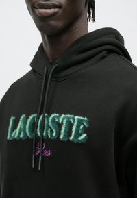 Czarna bluza z kapturem z puszystym zielonym logo „LACOSTE” oraz fioletowym napisem „Paris”. Sznurowadła są czarne z plastikowymi końcówkami. Miękka tekstura tkaniny.