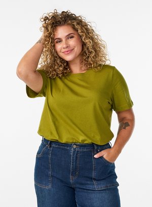 Krøllet-håret kvinde smiler med den ene hånd i lommen og den anden, der rører ved hendes hoved, iført en olivengrøn skjorte og blå jeans.
