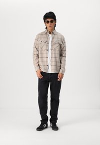 Lichtbeige geruite shirtjack met twee borstzakken, gedragen over een wit T-shirt, gecombineerd met zwarte jeans en zwarte sneakers.