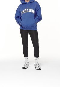 Modrá mikina s kapucí a grafikou "PEGADOR", v kombinaci s černými legínami a bílými sportovními botami s šedými akcenty.