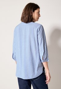 Chemise rayée bleu clair et blanche avec des manches trois-quarts et des fentes sur les côtés, présentant une coupe décontractée et un tissu texturé.