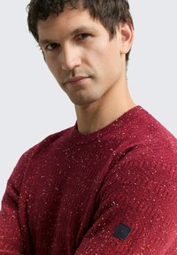 Pull en tricot bordeaux avec un motif texturé, col rond et manches courtes. Présente un petit logo de marque noir sur le bras gauche.