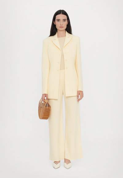 Cream-colored tailored suit with wide-legged trousers and a fitted blazer. Accessory includes a woven wooden handbag and pointed white heels.