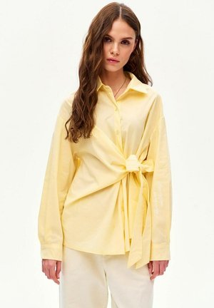 Kobieta o długich brązowych włosach, ubrana w jasnobrzoskwiniową bluzkę na guziki związaną w supeł z boku oraz kremowe spodnie na jednolitym tle.