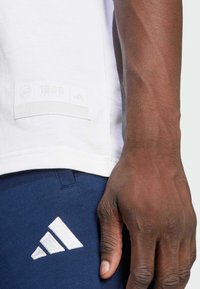 T-shirt blanc en tissu texturé, avec une étiquette cousue portant "1886 Arsenal" et le logo Adidas. Pantalon bleu foncé avec le logo blanc d'Adidas.