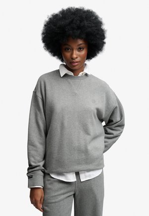 Kvinde med afro-frisure iført en grå sweatshirt og matchende bukser over en hvid skjorte med krave, stående med den ene hånd på hoften.