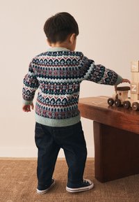 Flerfärgad stickad tröja med geometriska mönster och gröna detaljer, kombinerad med mörka jeans och svarta sneakers. Träleksaker på ett bord i närheten.