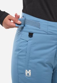 Un pantalon de ski bleu clair avec une taille réglable par velcro, des poches zippées et un logo de marque à l'avant. Texture de tissu lisse.