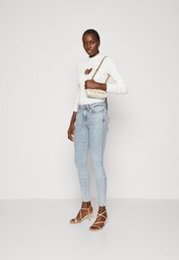 Suéter blanco de manga larga con corte en forma de corazón; jeans ajustados de lavado claro; tacones dorados con tiras; bolso pequeño con patrón y cadena.